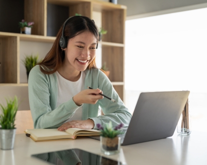 Picture of a girl studying on her laptop, wearing headphones and holding a pen in her hand