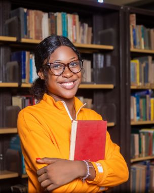 Portrait of a happy international student standing in library and holding a book in her arms