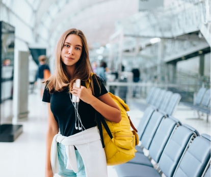 Picture of a girl standing at an airport, wearing bag on her shoulders and holding a phone in her hands